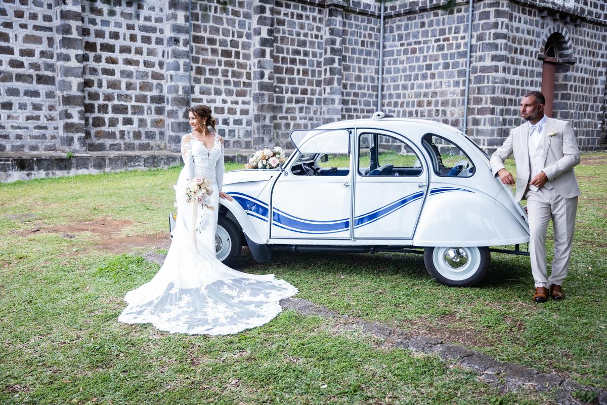 Emma et Fabien de part et d'autre de la 2CV fleurie devant l'église, la traîne de la robe déployée sur le sol