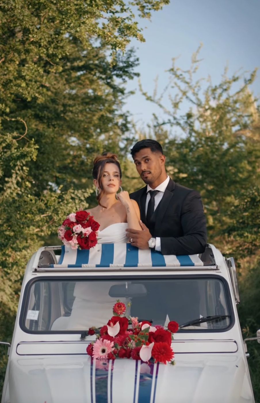 Des mariés debout dans la 2CV décorée de fleurs, au soleil couchant