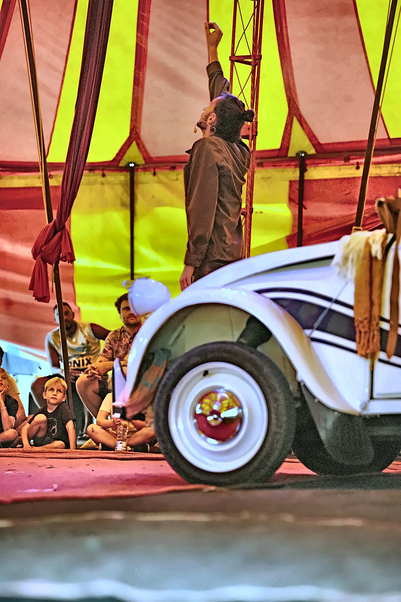 Artiste debout en équilibre sur le toit de la 2CV, parapluie levé, devant un public d'enfants